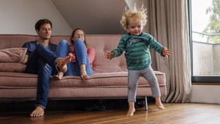 Ein Kind hüpft in die Luft, während ihm seine Eltern von einem Sofa aus zugucken.