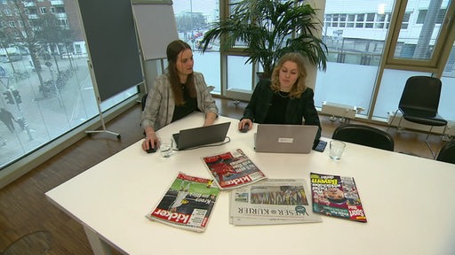Zwei Frauen sitzen vor Laptops an einem Tisch, auf dem Zeitungen und Magazine ausliegen.