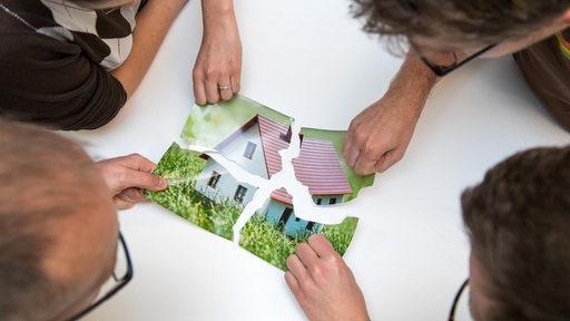 Vier Erwachsene schauen auf ein Bild von einem zerissenen Papierstück auf dem ein Haus zusehen ist