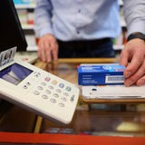 Die Versichertenkarte einer Krankenversicherung steckt in einem Kartenleseterminal, während ein Apotheker ein Medikament aushändigt. 