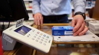 Die Versichertenkarte einer Krankenversicherung steckt in einem Kartenleseterminal, während ein Apotheker ein Medikament aushändigt. 