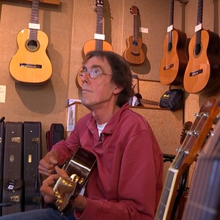 Ein Mann spielt Gitarre. Um ihn herum sind viele weitere Gitarren zu sehen.