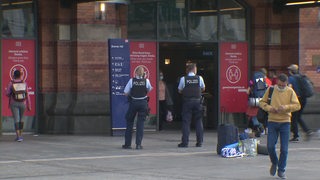 Zwei Polizisten stehen vor dem Haupteingang des Bremer Hauptbahnhofs. Passanten und Reisende laufen an ihnen vorbei.
