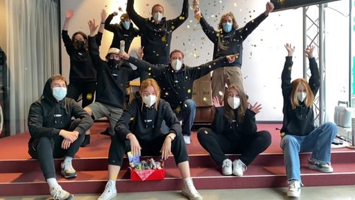 Mitarbeiter von Bremen Next sind für ein Foto aufgereiht und schmeißen Konfetti in die Luft. Alle tragen eine Maske.