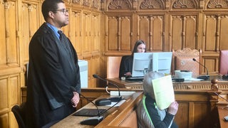 Angeklagter sitzt im Gerichtssaal und hält eine Akte vor sein Gesicht