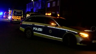 Ein Polizeifahrzeug steht nachts auf einer Straße. Im Hintergrund sind Krankenwagen zu sehen.