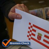 "Bremen-Check"-Logo vor einer Hand, die einen Brief mit Bremer Speckflagge in eine Urne wirft.