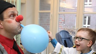 Ein Klinikclown bläst für einen kleinen Patienten einen Luftballon auf