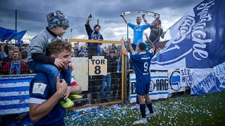 Torschütze Sebastian Kmiec vom Bremer SV läuft im Vordergrund entlang, im Hintergrund feiern Fans ihn und seine Mannschaft mit Fahnen und Schals.