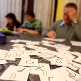 Auf einem Tisch liegen mehrere Karten mit Buchstaben, im Hintergrund schreiben Erwachsene auf Zettel.