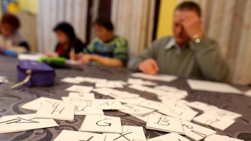 Auf einem Tisch liegen mehrere Karten mit Buchstaben, im Hintergrund schreiben Erwachsene auf Zettel.
