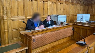 Zwei Männer sitzen vor einer Holzwand an einem Holztisch.