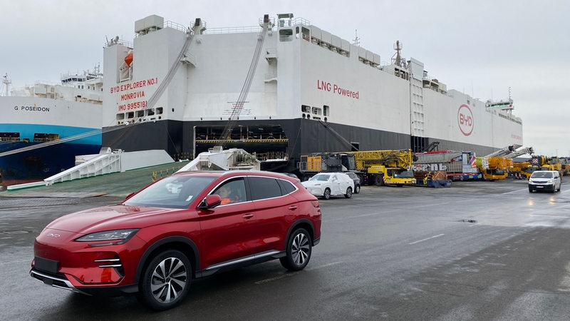 Vor einem im Hafen liegenden Autofrachter ist ein E-Auto geparkt.