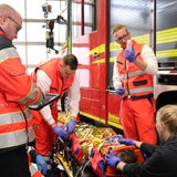 Mehrere Personen in Sanitäter-Kleidung stehen vor einem Feuerwehrauto um eine Person auf einer Trage herum.