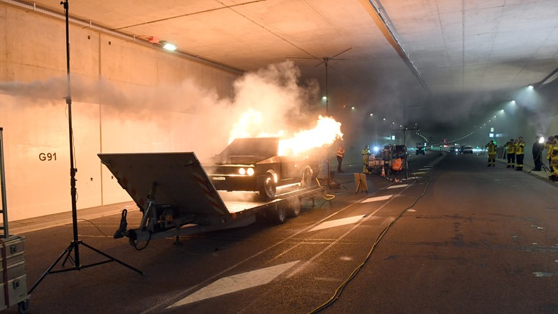 Eine Pkw-Attrappe steht in einem Tunnel in Flammen, drumherum stehen Feuerwehrleute.