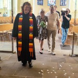 Ein Brautpaar tritt hinter einer Pastorin vor Zuschauer
