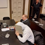 Zwei Personen sitzen an Tischen und halten sich Ordner vor das Gesicht. Daneben sitzen Personen in schwarzen Roben.