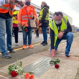 Ein Mann kniet vor einer Gedenktafel neben der Rosen liegen, drumherum stehen Menschen in Warnwesten.