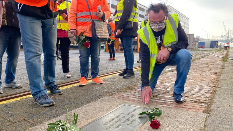 Ein Mann kniet vor einer Gedenktafel neben der Rosen liegen, drumherum stehen Menschen in Warnwesten.