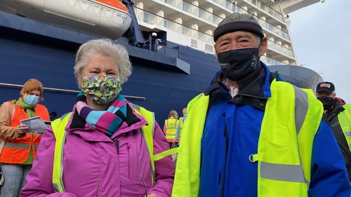 Ein älteres Ehepaar mit Warnweste und Mundschutz steht vor einem Kreuzfahrtschiff.