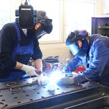 Zwei Männer in blauer Arbeitskleidung stehen an einem Tisch und schweißen.
