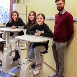 In einer Schule in Bremerhaven sitzen zwei Schülerinnen auf Deskbikes. Zwischen ihnen steht eine Mitschülerin, hinter ihnen lehnt ein Lehrer an der Wand.