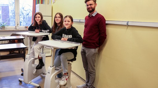 In einer Schule in Bremerhaven sitzen zwei Schülerinnen auf Deskbikes. Zwischen ihnen steht eine Mitschülerin, hinter ihnen lehnt ein Lehrer an der Wand.