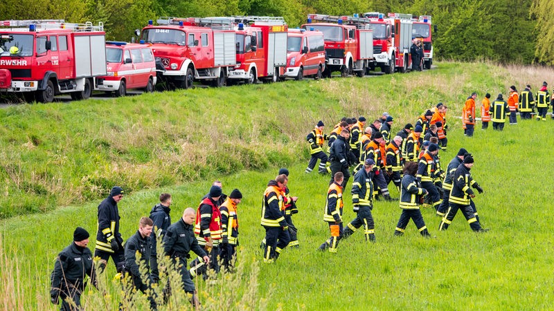 Feuerwehrleute gehen in einer breiten Reihe eine Wiese entlang. Im Hintergrund stehen Feuerwehrautos auf einem Deich.