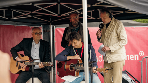 Vier Männer stehen unter einem Pavillon. Zwei spielen eine Gitarre und die anderen zwei stehen dahinter. Einer hält ein Mikro