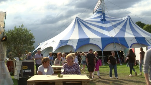 Auf der Breminale ist ein großes blau-weiß gestreiftes Zelt zu sehen. Im Vordergrund sitzen und laufen Personen.