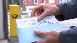 Ein Muster von einem Stimmzettel in einem Briefumschlag wird vor einen Postkasten gehalten. 