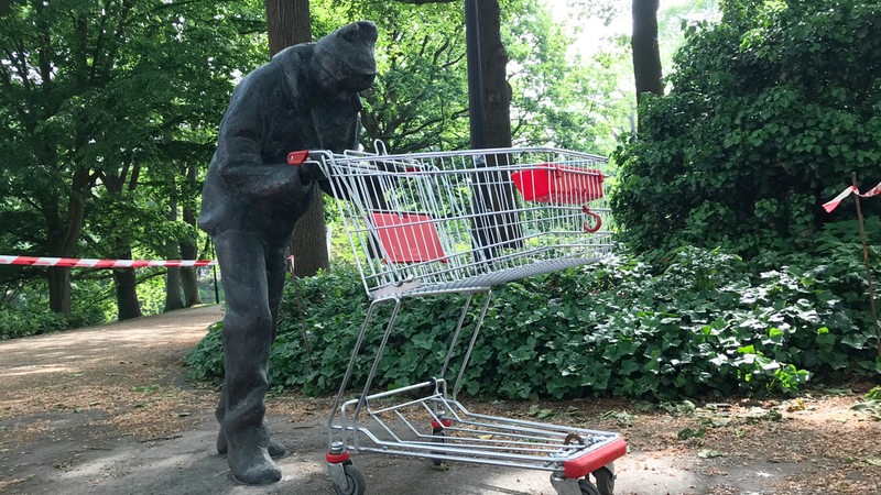 Ein Mann aus Bronze schiebt einen echten Einkaufswagen