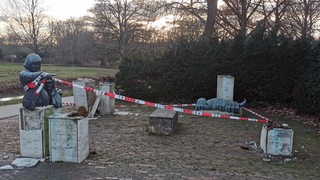 Eine Bronzefigur auf einem Sockel. Im Hintergrund eine liegende Bronzefigur. Daneben mehrere leere teils zerstörte Betonsockel. Absperrband. 