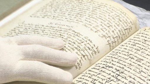 Zu sehen ist ein aufgeschlagenes Buch und eine Hand mit Handschuh, die dieses Umblättert.