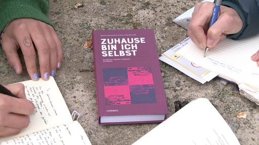 Ein Buch in der Mitte, auf dem "ZUHAUSE BIN ICH SELBST" steht. Neben dem Buch zwei offene Notizhäfte, in denen geschrieben wird.