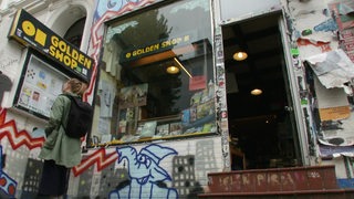 Vor dem Laden Golden Shop steht ein Frau und liest in einem Schaukasten. Die Fassade ist mit bunten Graffitis verzieht.