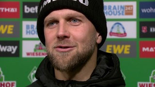 Werder-Stürmer Niclas Füllkrug mit einer schwarzen Wollmütze beim Interview vor einer Werbewand.