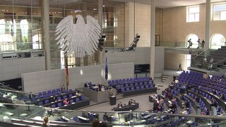 Der deutsche Bundestag von innen. Blaue Stühle sind in einem Halbkreis um ein Rednerpult angeordnet. 