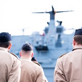 Soldaten stehen im Marinestützpunkt Wilhelmshaven mit dem Rücken zur Kamera. Im Hintergrund ist ein Schiff zu sehen.