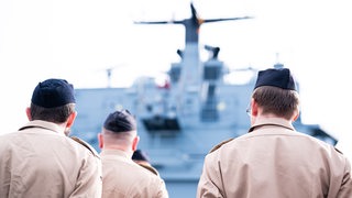 Soldaten stehen im Marinestützpunkt Wilhelmshaven mit dem Rücken zur Kamera. Im Hintergrund ist ein Schiff zu sehen.