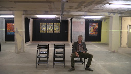 In einem Bunker sieht man im Hintergrund Gemälde an den Wänden hängen. Im Vordergrund stehen Stühle und auf einem Sitz ein Mann.
