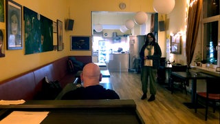Ein Kunde sitzt in einem Café und wird von einer Frau mit Schutzmaske bedient.