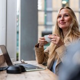 Eine blonde Frau sitzt in einem Café vor ihrem Laptop, hält eine Kaffeetasse in der Hand und schaut in die Ferne.