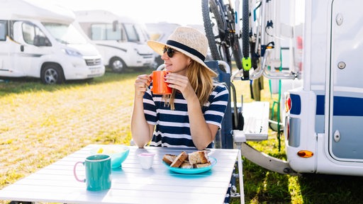 Eine Frau mit Hut und Sonnenbrille bei einem Frühstück auf einem Campingplatz (Symbolbild)
