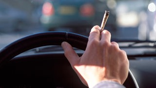 Eine Person sitzt am Steuer eines Autos und hält einen Joint in der Hand.
