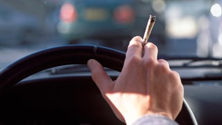 Eine Person sitzt am Steuer eines Autos und hält einen Joint in der Hand.