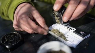 Ein Mann schüttet zerkleinertes Marihuana in einen Joint. Zu sehen sind nur seine Finger, Tabakblättchen und eine Dose.