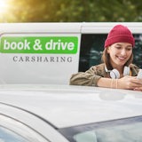 Ein junge Frau bucht mit einem Mobiltelefon ein Auto. Im Hintergrund ein Bus mit der Aufschrift "book & drive - Carsharing"
