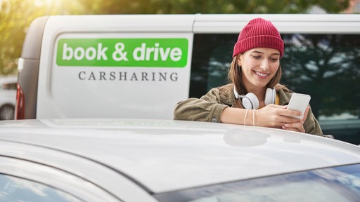 Ein junge Frau bucht mit einem Mobiltelefon ein Auto. Im Hintergrund ein Bus mit der Aufschrift "book & drive - Carsharing"