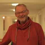 Ein Mann lächelt in die Kamera, er trägt einen roten Pullover, eine schwarze Brille, einen grauen Dreitagebart und eine graue Kurzhaarfrisur 